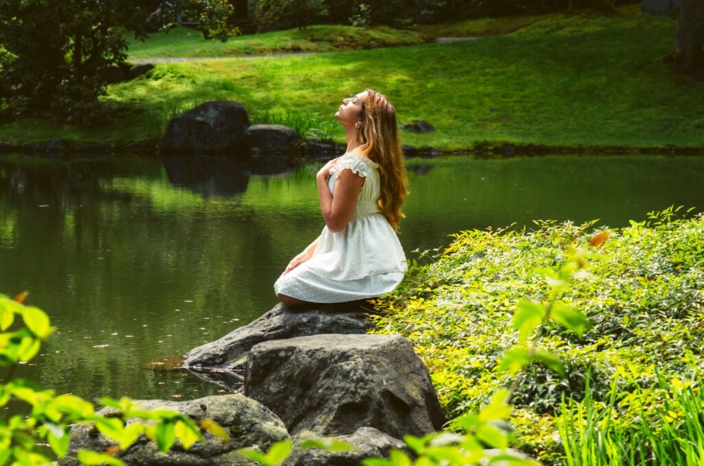 A woman meditates by a tranquil river in Vancouver, BC, surrounded by lush greenery.