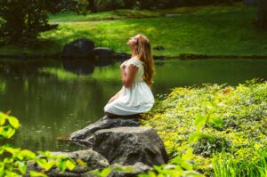 A woman meditates by a tranquil river in Vancouver, BC, surrounded by lush greenery.