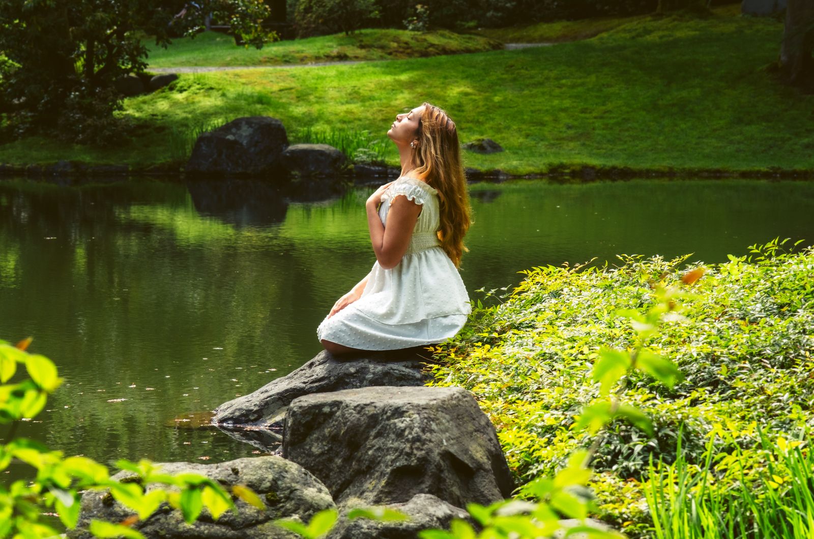 A woman meditates by a tranquil river in Vancouver, BC, surrounded by lush greenery.
